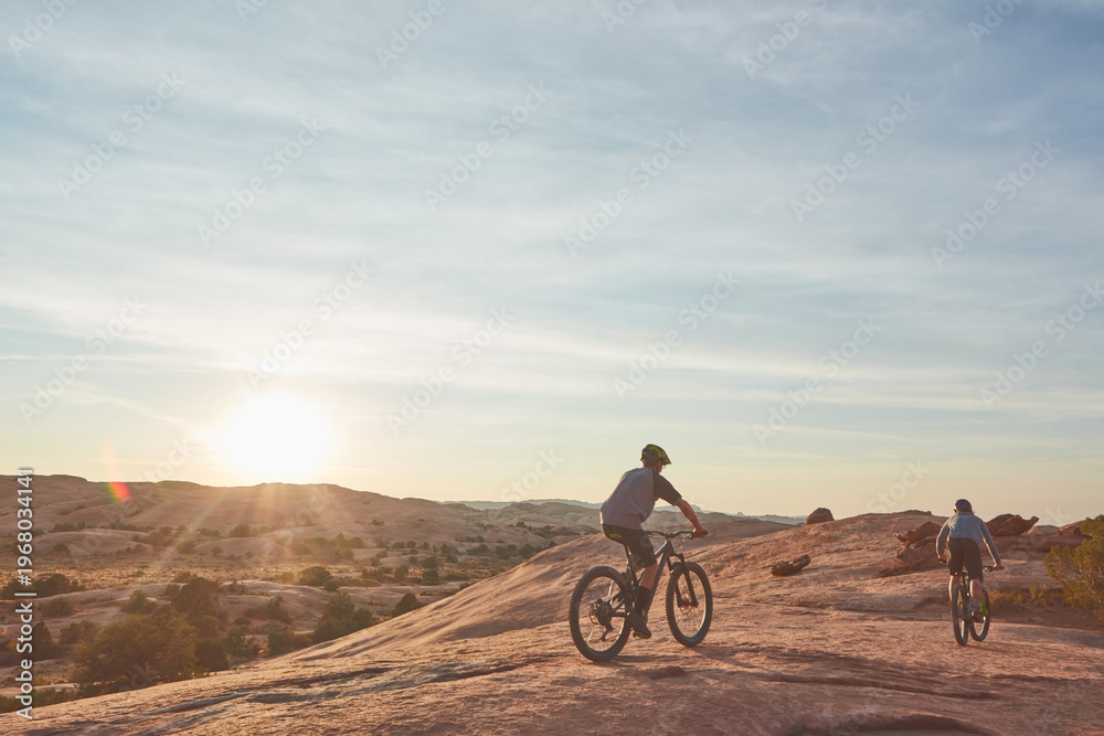 Fototapeta premium Men, mountain bike or cycling with sunset in desert or dunes for extreme challenge or sport. Active, male people or cyclist with bicycle for outdoor trip or travel on dirt track together in nature