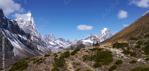 Breathtaking wide panoramic view of snow-capped Himalayan range under bright sunny sky, with two trekkers on Everest Base Camp trail - one riding horse with backpack high mountain path 