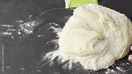Female baker kneads dough on a black countertop, hands covered in flour, with scattered flour and a green spatula visible in the kitchen setting
