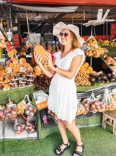 Stylish woman enjoying shopping at tropical fruit stall, holding ripe pineapple. Bright summer mood, exotic products and authentic local market experience during vacation
