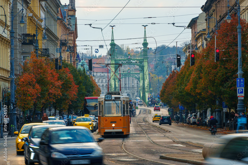 Obraz premium Yellow retro tram riding through the autumn street of Budapest toward Liberty Bridge with red trees.