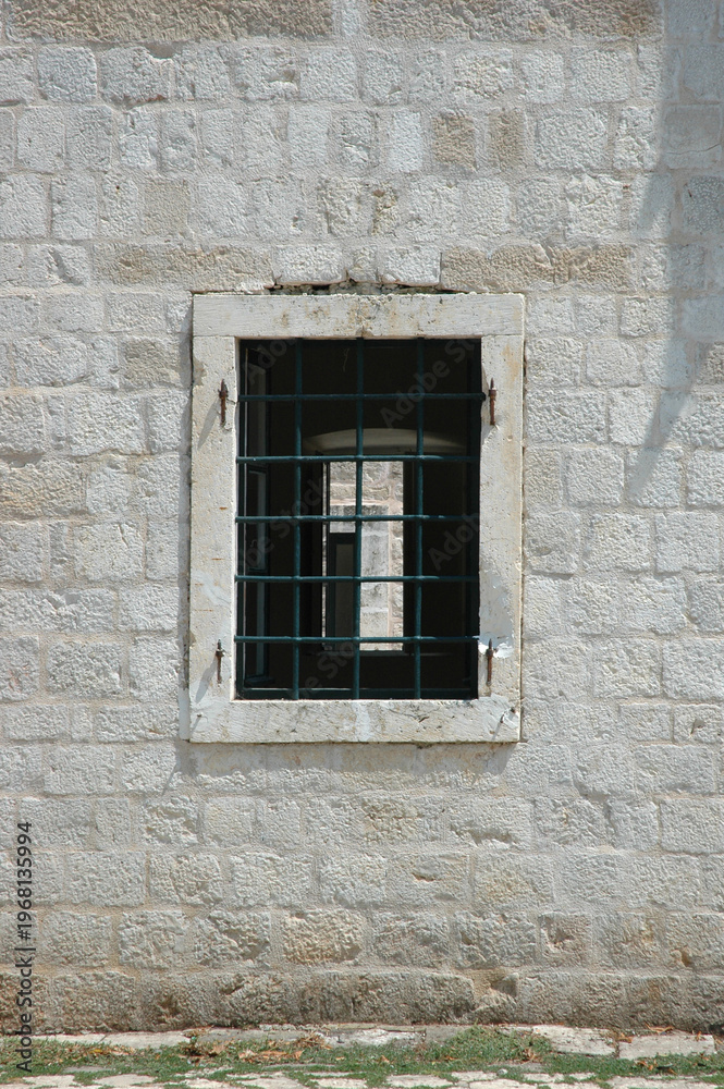 Fototapeta premium A traditional window with a white stone frame and green iron bars is set into a weathered light stone wall in the town of Ston