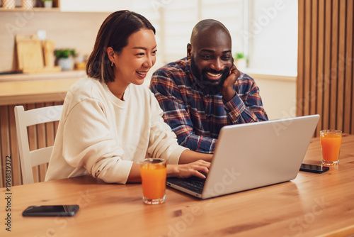 A cheerful couple sits together at a wooden table, smiling and laughing while looking at a laptop screen in a bright and cozy home kitchen.