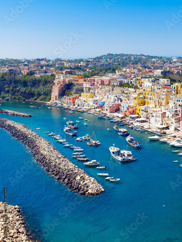 Marina Corricella harbor on Procida with colorful houses along waterfront. Procida coastline with breakwater, moored boats and clear Mediterranean water under bright sky in Campania region