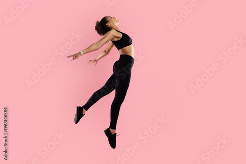Tableau sur toile Athletic black woman jumping backward with arms open on pink studio background,
