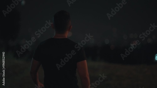 Cinematic footage of a man standing alone overlooking city lights at night. Urban night scene with soft bokeh lights, representing solitude, reflection, and emotional mood in a modern city environment