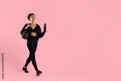 Young sporty black woman in black activewear walking confidently, holding water bottle and gym bag, smiling on pink studio background. Copy space