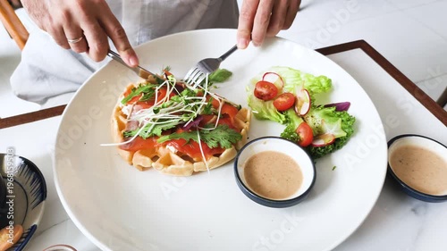 Hands cut into healthy breakfast at table. Person uses knife for waffle with salmon. High angle shows healthy breakfast with salad. Fresh meal includes sauce. Nutritious food is served.