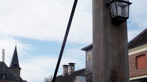 Historic brick buildings with a clock tower and lamp post are shown outdoors under a partly cloudy sky in a quiet street setting.