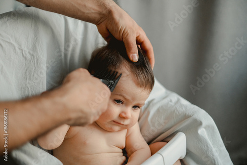 Soft light falls on a baby's gentle face. Crisp hands brush through fine, dark hair with care. Baby gazes curiously, eyes wide and trusting. Calm moment of tender care, intimate and quiet
