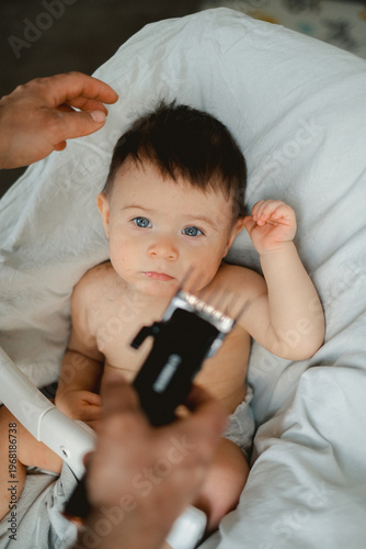 Soft light catches a baby's curious gaze. Hands gently guide a trimmer near the child's head. Baby's expression is calm, slightly wide-eyed. Tender moment during a gentle haircut