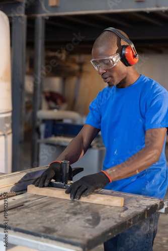 Table saw slicing wooden plank on workshop workbench, showing dust-covered tools and beams overhead