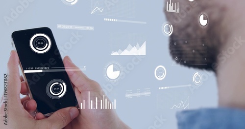 Holding smartphone, bearded man wearing blue collared shirt studying floating HUD graphs at office