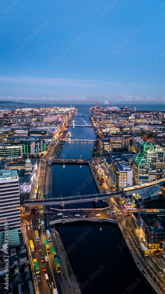 Fototapeta premium Portrait shot looking down river Liffey in Dublin at blue hour dawn night time with illuminated cityscape buildings and clear blue sky in winter