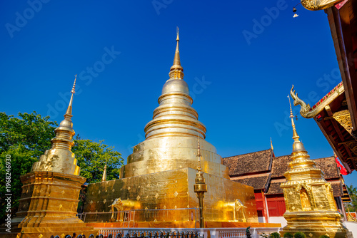 The grand golden chedi (stupa) of Wat Phra Singh Woramahavihan, featuring three main stupas and elaborate Lanna architecture against a vibrant blue sky in Chiang Mai, Thailand.