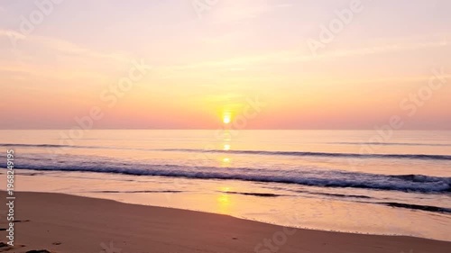 The sun rises over a calm ocean, casting a warm golden glow across the tranquil water and sandy shore