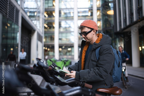 Photography Young man in beanie and sunglasses checks his smartphone beside a row of shared