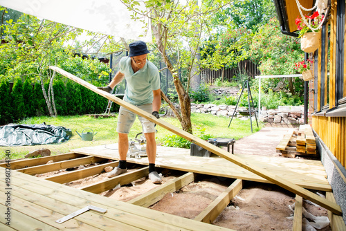 Man building wooden deck in backyard. Authentic DIY home improvement project during summer. Outdoor house renovation and construction.