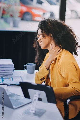 Businesswoman experiencing discomfort and pain in her lower back while sitting at her desk, working on a laptop in a modern office environment, highlighting work-related health issues and stress