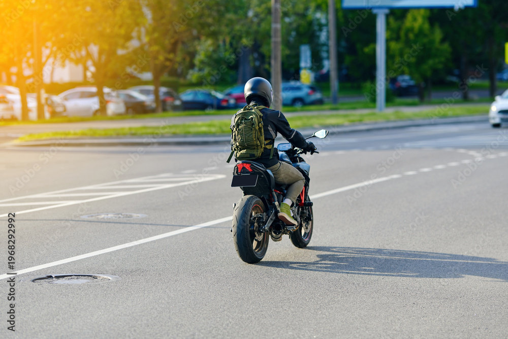 Obraz premium Motorcycle with rider in protective gear and an olive green bag passing through an intersection during sunny day. Biker on black sport motorcycle, sportbike in motion, rider wearing protective helmet