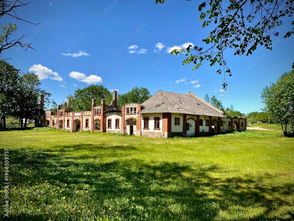 Obraz premium An abandoned historic palace with red brick and plaster walls stands in a grassy field under a clear blue sky. The building displays a series of arches and large, arched windows