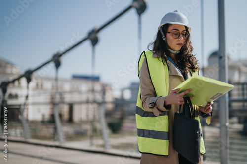 Obraz na płótnie A female engineer in a hard hat and high-visibility vest reviews documents on a bridge