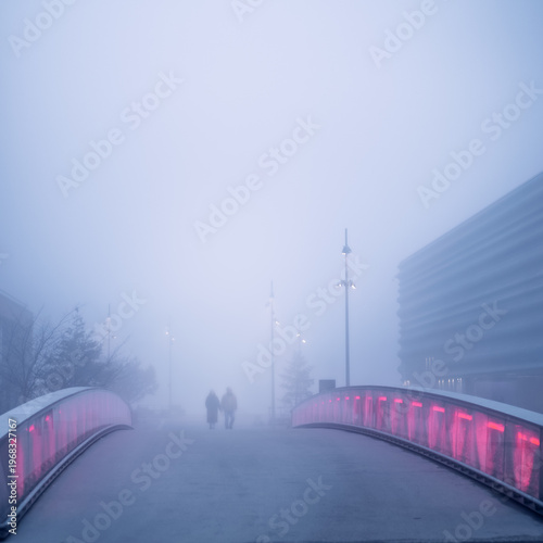 Couple in fog crossing a bridge with red lights in guardrails.