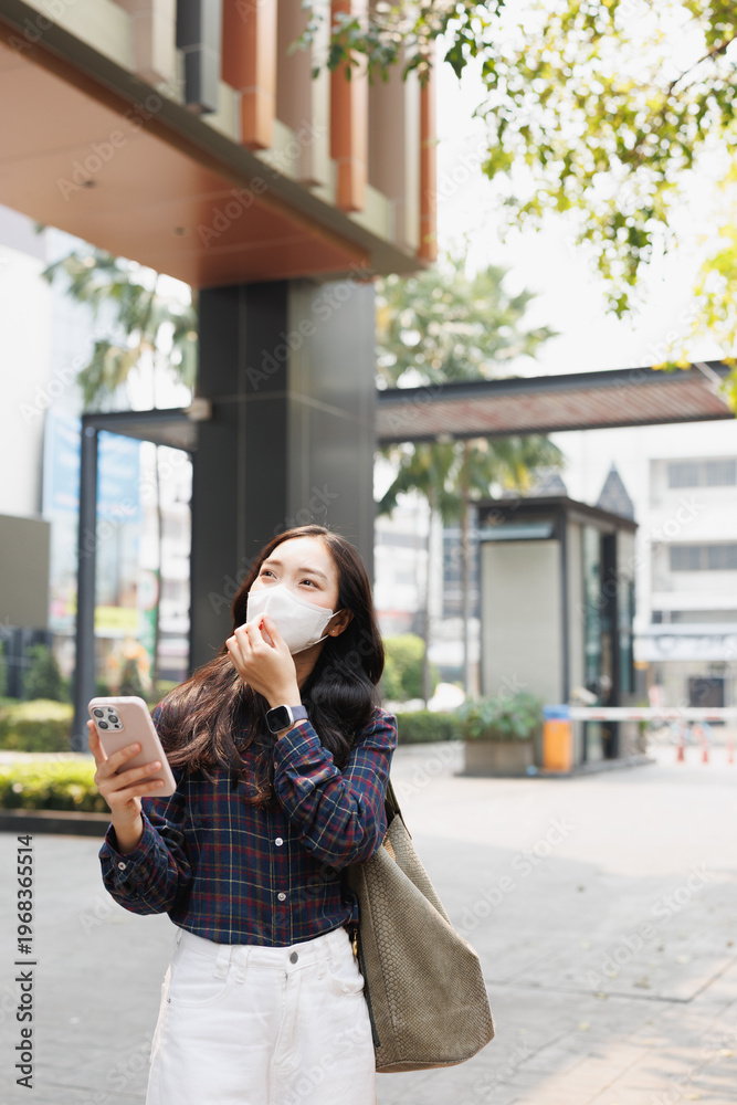 Fototapeta premium Young woman confidently walking in an urban environment while wearing a face mask for protection and holding a modern smartphone, representing changing city lifestyles