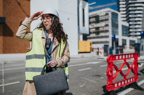 Vászonkép A female construction engineer wearing a hard hat and hi vis vest holds a briefcase at a city construction site