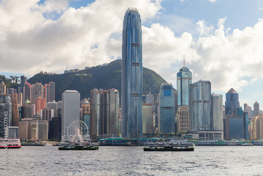 Fototapeta premium Hong Kong skyline dominated by the International Finance Centre, ferries crossing Victoria Harbour and a bustling waterfront of high-rise buildings against green hills and dramatic clouds