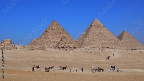 Giza pyramids with camels, horses and carriages crossing sandy desert plateau, Egypt. Iconic ancient wonder under vivid blue sky. Travel and history.