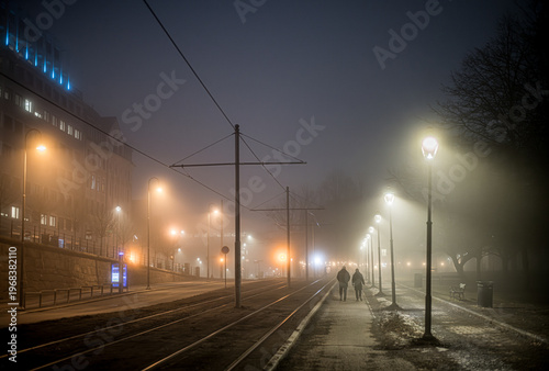 Two people walking along a lamp-lit pavement a foggy evening