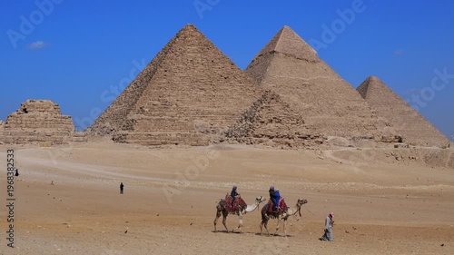 Giza pyramids with camels, horses and carriages crossing sandy desert plateau, Egypt. Iconic ancient wonder under vivid blue sky. Travel and history.