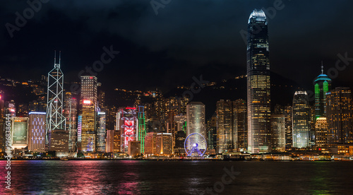 A panoramic night view of Hong Kong skyline across Victoria Harbour, featuring illuminated skyscrapers, colorful waterfront lights and reflections on the water