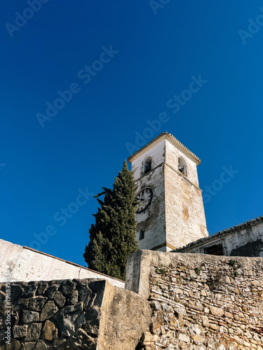 Old white church tower behind a stone wall under a clear blue sky. Colmenar, Malaga, Andalusia, Spain.
