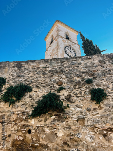 Old white church tower behind a stone wall under a clear blue sky. Colmenar, Malaga, Andalusia, Spain.