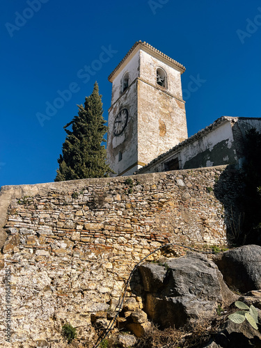 Old white church tower behind a stone wall under a clear blue sky. Colmenar, Malaga, Andalusia, Spain.