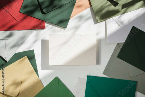 Overhead view of a paper card mockup over assorted multi coloured envelopes on a table in dappled light.