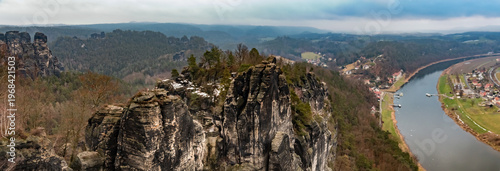 Large panorama from the Basteiaussicht with the jagged Bastei rock formations centered, sandstone cliffs to the left, and the Elbe Valley with the river sweeping along the right on a cold winter day.