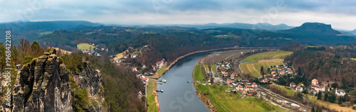 Large winter panorama of the Elbe Valley with the village of Kurort Rathen nestled along the river, framed by the towering Bastei rock formations rising on the left. Captured from the Basteiaussicht.