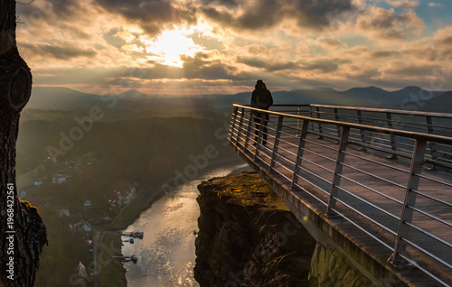 Stunning photo of a man standing alone on the popular viewing platform Basteiaussicht, admiring the panoramic view over the Elbe Valley during a cold winter sunrise, with the sun in front of him.