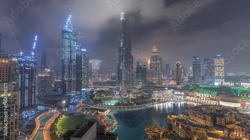 Bright illuminated modern skyscrapers rising above the famous mall and fountain in Dubai Downtown district. Aerial panoramic night view under a cloudy sky in time-lapse.