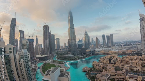 Tall skyscrapers rising above the famous mall and fountain in Dubai Downtown district. Aerial panoramic top view showing day to night transition with cloudy sunset sky.