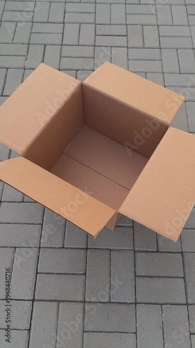 An empty open cardboard box sitting on a grey paved outdoor surface with its flaps fluttering in the wind. Close up.