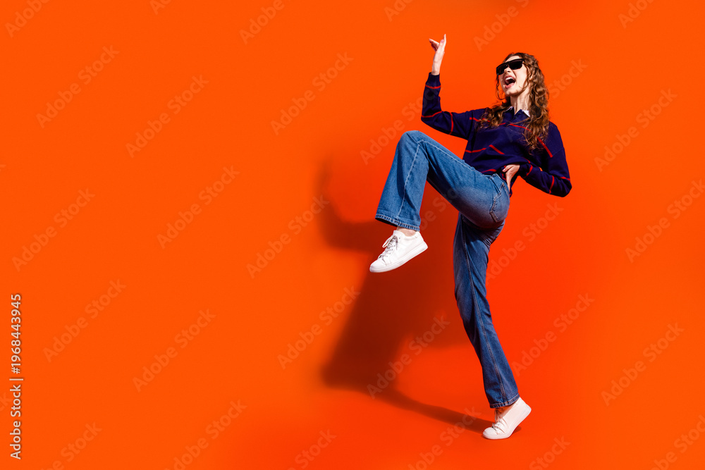Obraz premium Stylish woman in sunglasses dancing energetically against a vibrant orange background wearing jeans and white sneakers