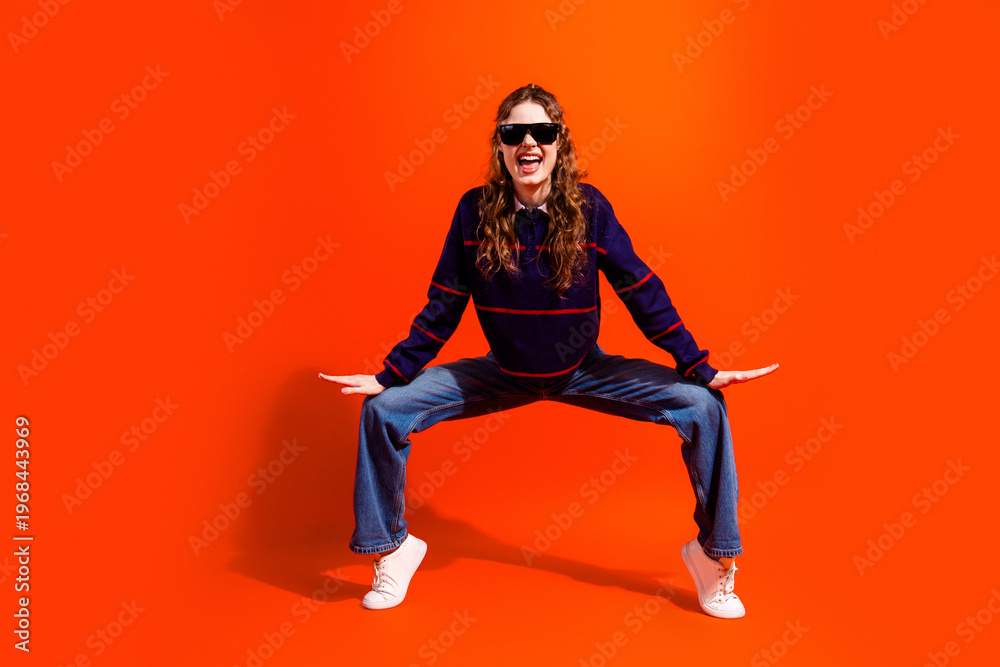Obraz premium Young woman in sunglasses striking an energetic wide-legged pose wearing denim jeans and sneakers against a bold orange background