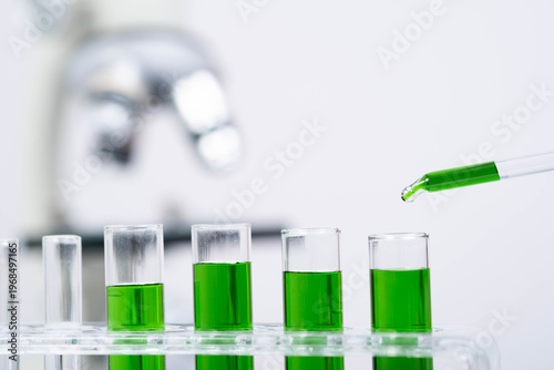 Pipette dropping green chemical liquid into laboratory test tubes with blurred microscope background. Biotechnology research, chemistry experiment, microbiology testing, pharmaceutical science concept