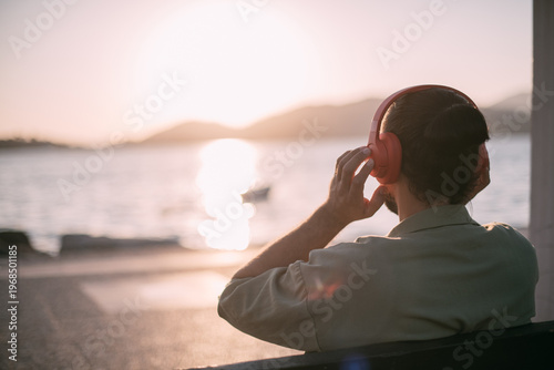 Portrait from the back of a young man in bright big headphones by the sea at sunset. A handsome guy listens to music on the ocean in the rays of the sun, with his back to the camera