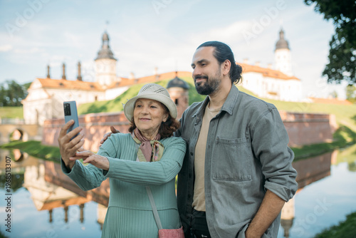 A pensioner and her adult son on a guided tour of an ancient castle. They smile happily and take selfies on their phones. An elderly mother and son travel together, visiting historical sites