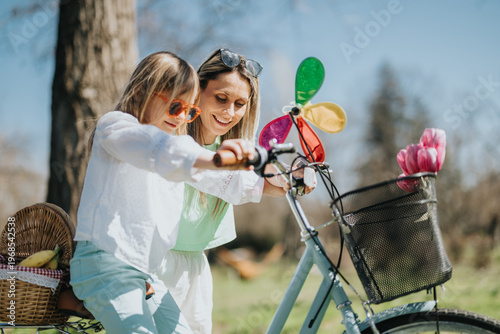 A mother helps her young daughter ride a bicycle while smiling and guiding her hands. The pair practice together outdoors beside a picnic basket and flowers on a sunny day.
